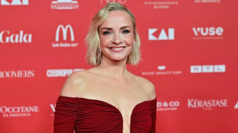 Janin Ullmann Janin Ullmann steht vor einer Pressewand. Sie trägt ein dunkelrotes, schulterfreies Kleid mit tiefem Ausschnitt und lächelt in die Kamera.