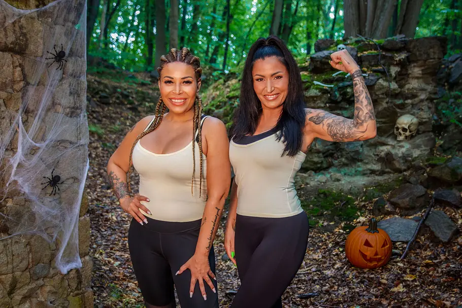 Jasmin Herren & Patricia Blanco Jasmin Herren & Patricia Blanco posieren zwischen Halloween-Dekorationen im Wald. Eine winkelt ihren tätowierten Arm nach oben an.