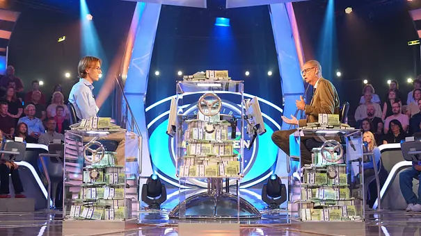 Günther Jauch und Kandidat Marco sitzen in einem Fernsehstudio an transparenten Pulten mit Geldbündeln. Dahinter sitzt Publikum im blauen Licht. - Foto: RTL / Stefan Gregorowius Günther Jauch und Kandidat Marco sitzen in einem Fernsehstudio an transparenten Pulten mit Geldbündeln. Dahinter sitzt Publikum im blauen Licht. - Foto: RTL / Stefan Gregorowius