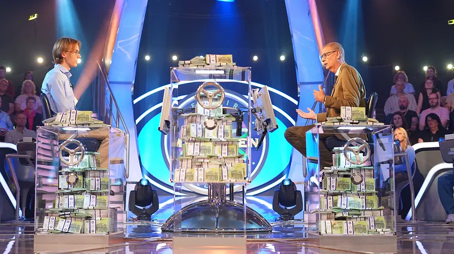 Günther Jauch hilft Kandidat Marco bei einer Frage auf die Sprünge. Günther Jauch und Kandidat Marco sitzen in einem Fernsehstudio an transparenten Pulten mit Geldbündeln. Dahinter sitzt Publikum im blauen Licht.