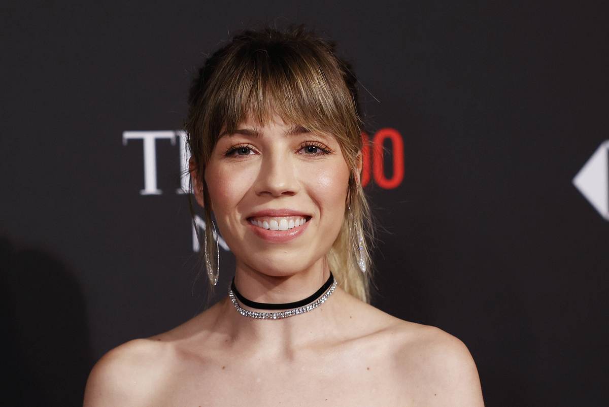 Jennette McCurdy lächelt vor einer Fotowand. Sie trägt Ohrringe und eine Halskette, sowie ein schulterfreies Outfit. 