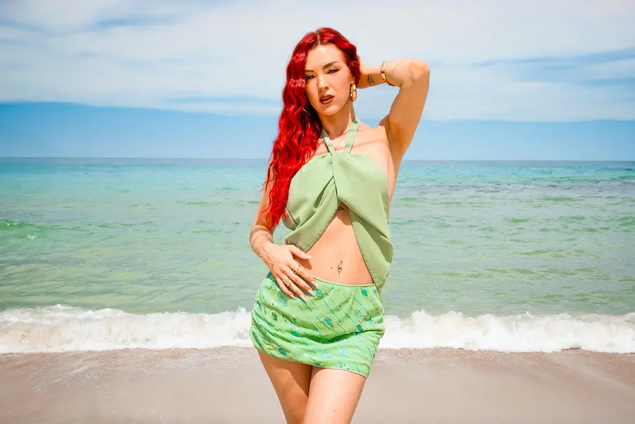 Jennifer Degenhart Jennifer Degenhart steht am Strand vor türkisblauem Meer. Sie trägt ein hellgrünes, bauchfreies Oberteil mit gekreuztem Schnitt und einen passenden Minirock, während hinter ihr sanfte Wellen an den Sand rollen.