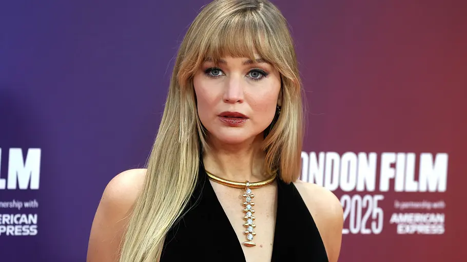 Jennifer Lawrence Brust OP Jennifer Lawrence trägt ein schwarzes Kleid und eine goldene Halskette vor einer violett-roten Wand mit Festival-Schriftzügen.