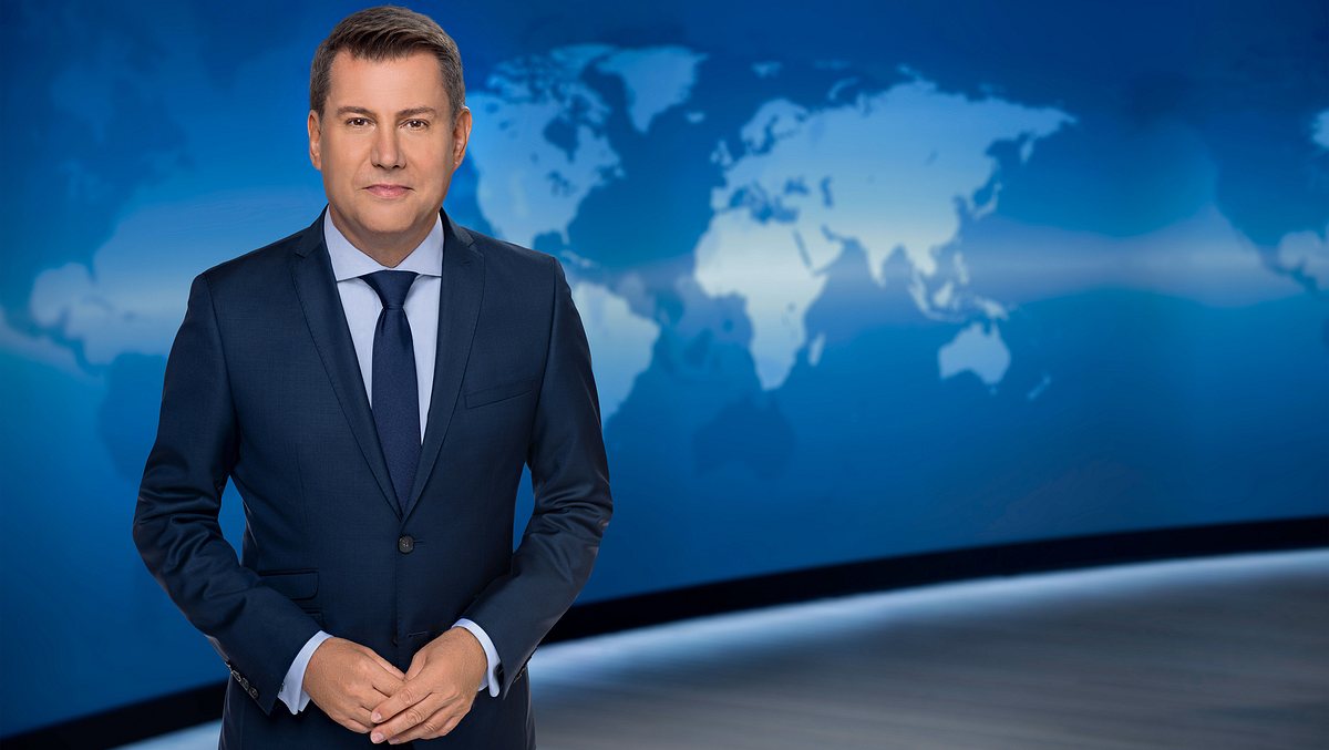 Jens Riewa im „Tagesschau“-Studio. Jens Riewa steht im dunkelblauen Anzug im „Tagesschau“-Studio. Er schaut in die Kamera und lächelt leicht.