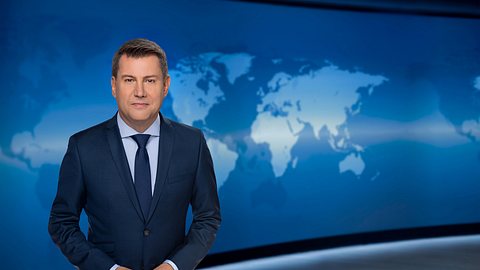 Jens Riewa steht im dunkelblauen Anzug im „Tagesschau“-Studio. Er schaut in die Kamera und lächelt leicht. - Foto: © NDR/Thorsten Jander