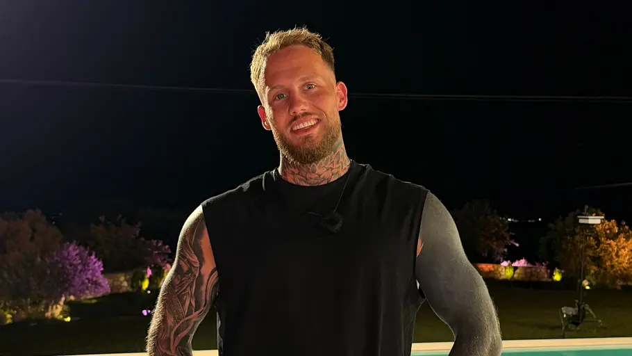 Jerry, 31 Ein tätowierter Mann in schwarzem ärmellosem Shirt steht nachts mit den Händen in den Hosentaschen vor einem beleuchteten Pool.