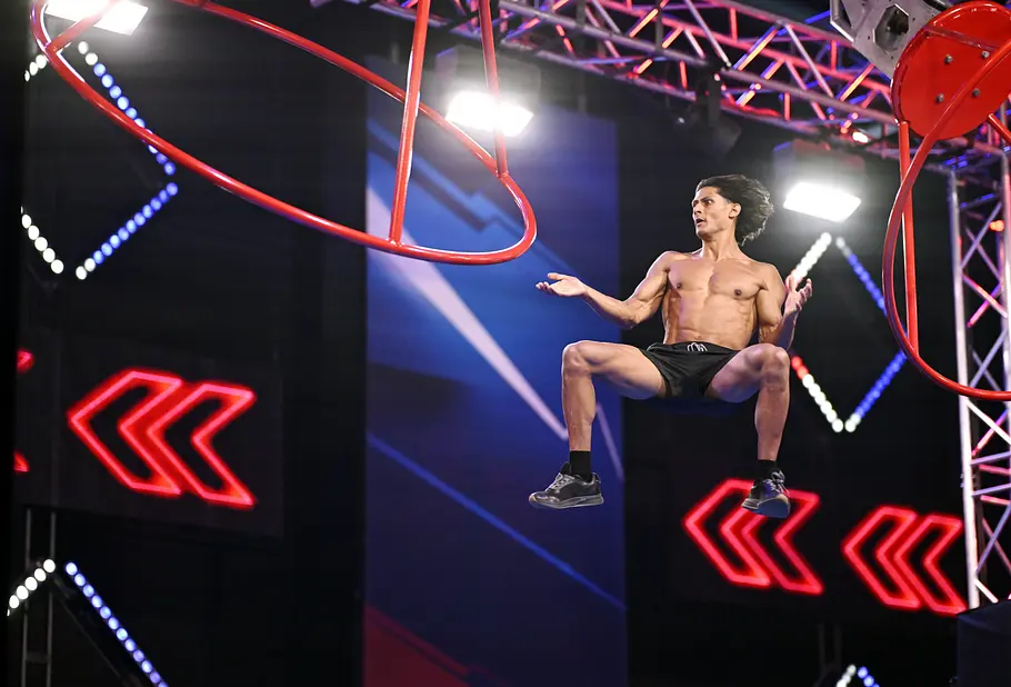 Jescher Heidl ist Teilnehmer bei Ninja Warrior Germany 2026 Jescher Heidl springt mit freiem Oberkörper und angewinkelten Beinen zwischen roten Hindernissen im beleuchteten Parcours.
