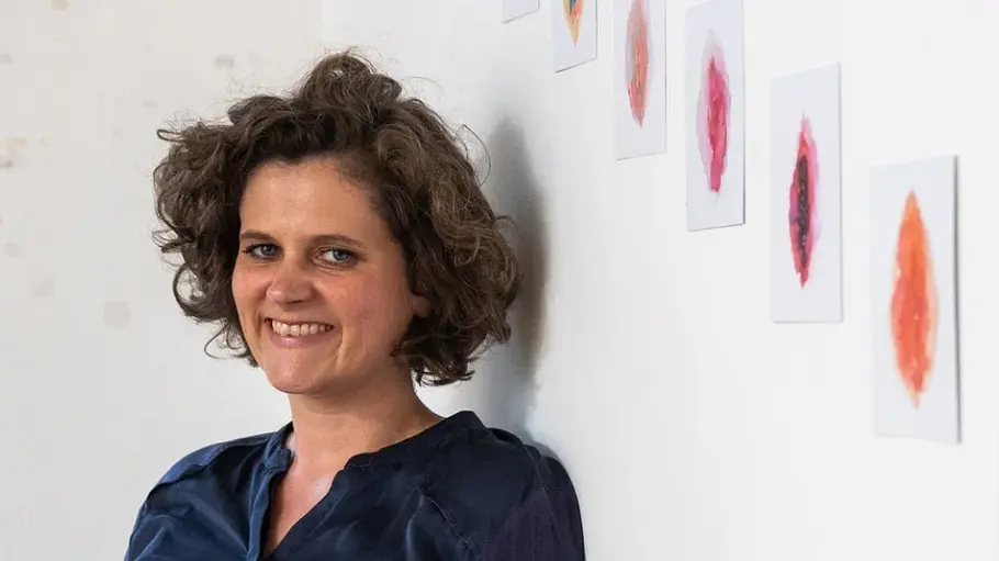 Jill Tammling Eine lächelnde Frau mit lockigem Haar und dunkelblauer Bluse lehnt an einer Wand mit bunten Vulva-Zeichnungen.