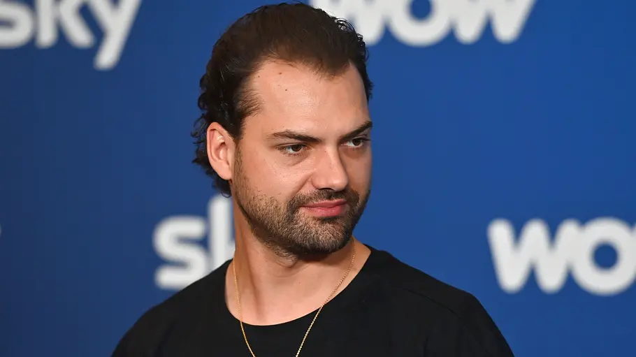 Jimi Blue Ochsenknecht will ins Dschungelcamp Jimi Blue Ochsenknecht trägt ein schwarzes Shirt und eine goldene Kette vor einer blauen Wand mit Sky- und WOW-Logos.