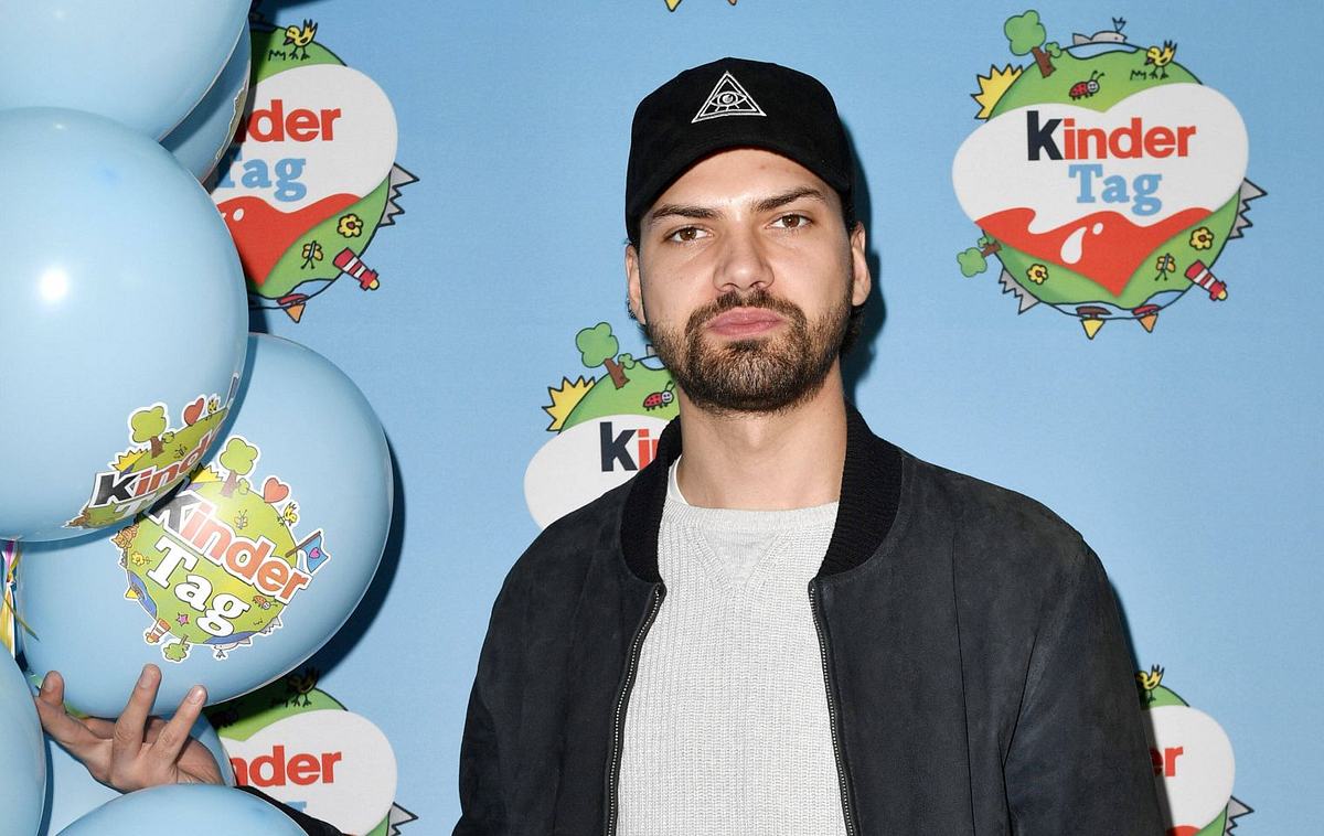 Jimi Blue Ochsenknecht Man sieht Jimi Blue Ochsenknecht vor einem Backdrop des Kinder-Tages. Er trägt einen grauen Pulli, eine schwarze Jacke und eine schwarze Basecap.
