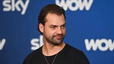 Jimi Blue Ochsenknecht trägt ein schwarzes Shirt und eine goldene Kette vor einer blauen Wand mit Sky- und WOW-Logos. - Foto: IMAGO / Sven Simon