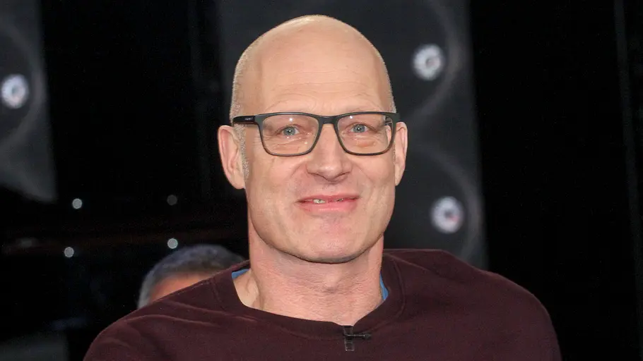 Joachim Meyerhoff Joachim Meyerhoff trägt eine schwarze Brille und einen dunkelroten Pullover. Er lächelt vor dunklem Hintergrund.