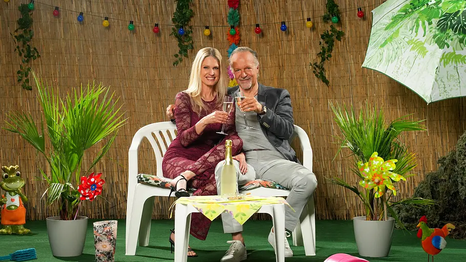 Jochen und Tina Horst Jochen und Tina Horst sitzen mit Sektgläsern auf einer weißen Gartenbank zwischen Topfpalmen und bunter Gartendekoration.