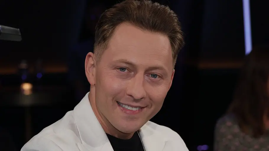 Jodok Cello Jodok Cello sitzt im Sessel im Studio der „NDR Talk Show“ und lächelt in die Kamera. Er trägt ein weißes Sakko über einem schwarzen Shirt mit Kette; im Hintergrund ist das Publikum unscharf zu sehen