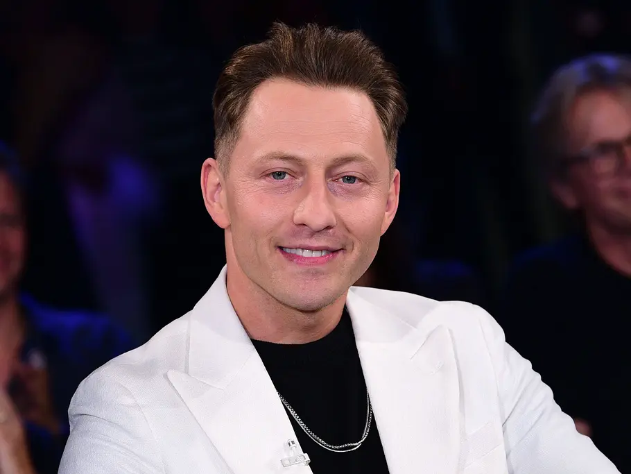 Jodok Cello lächelt im weißen Sakko über schwarzem Oberteil. Im dunklen Hintergrund sitzen unscharf weitere Personen.