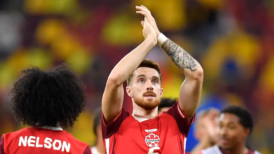 Ein kanadischer Nationalspieler mit der Rückennummer 5 applaudiert nach dem Spiel mit erhobenen Händen den Fans. Im Vordergrund ist ein weiterer Spieler mit der Rückennummer 21 von hinten zu sehen, während im Hintergrund weitere Teamkollegen und die unscharfen Tribünen eines Stadions erkennbar sind.
