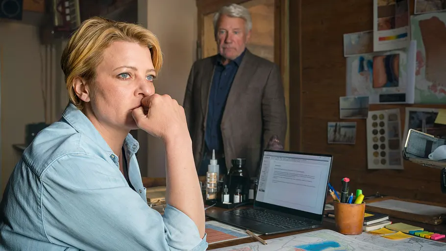 Jördis Triebel Neues Land Jördis Triebel sitzt mit der Hand am Mund vor Laptop und Landkarte am Schreibtisch. Hinter ihr steht ein älterer Mann im Sakko.