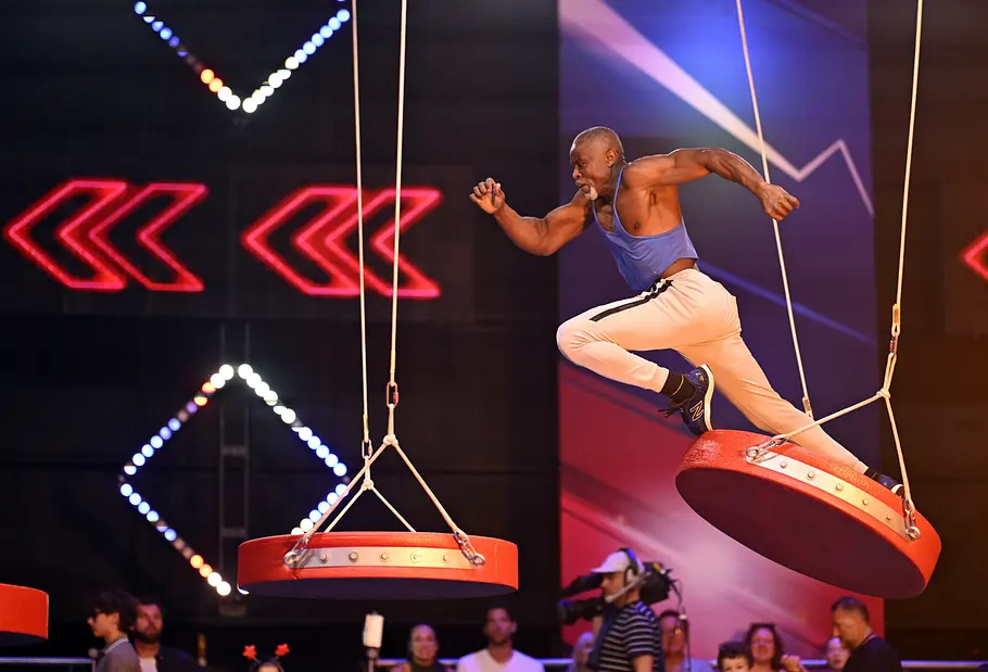 Joh-Anani Kpalete ist Teilnehmer bei Ninja Warrior Germany 2026 Joh-Anani Kpalete springt in blauem Oberteil und weißer Hose zwischen roten, an Seilen hängenden Plattformen.
