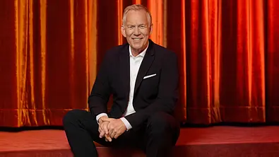 Johannes B. Kerner sitzt lächelnd in dunklem Anzug und weißen Turnschuhen auf einer roten Stufe vor einem orangefarbenen Vorhang. - Foto: ZDF und Christoph Neumann Johannes B. Kerner sitzt lächelnd in dunklem Anzug und weißen Turnschuhen auf einer roten Stufe vor einem orangefarbenen Vorhang. - Foto: ZDF und Christoph Neumann