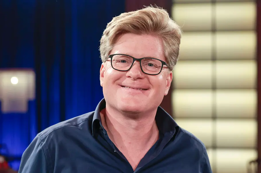 Johannes Schröder Johannes Schröder lächelt mit schwarzer Brille und dunkelblauem Hemd vor einem blauen Vorhang und hellen Leuchtflächen.