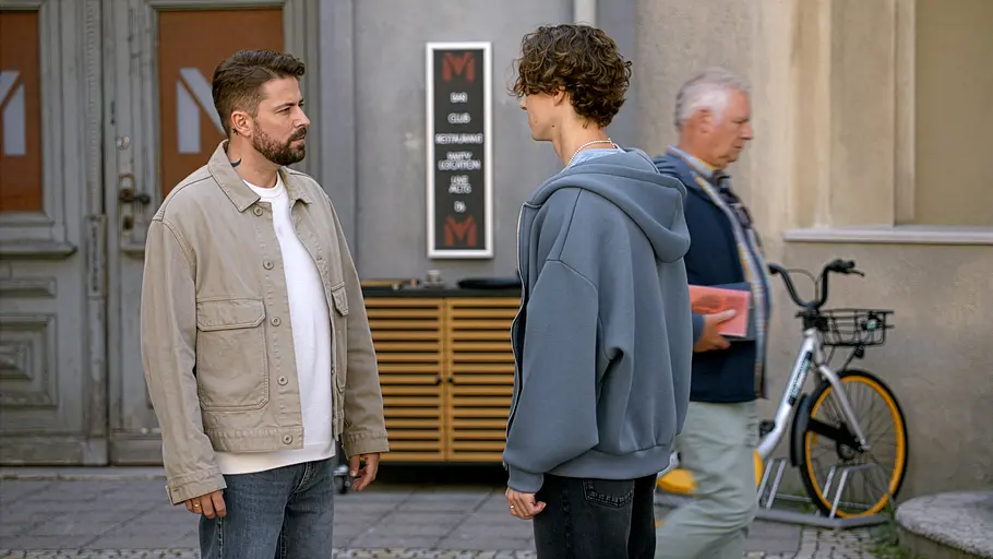 John steht links in beiger Jacke Benni im blauen Hoodie gegenüber. Im Hintergrund sind ein Passant und ein Fahrrad zu sehen.