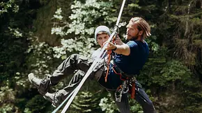 Die Bergretter: Ohne Ausweg: Im Wald: Über einem Abgrund ist eine Slackline gespannt. An dieser hängt Jonas Maier (Juls Serger) in seinem gesicherten Klettergurt und wirkt benommen. Vor ihm, ebenfalls gesichert, hängt Markus Kofler (Sebastian Ströbel) und zieht sich und Jonas an der Slackline entlang. - Foto: ZDF/Sabine Finger Fotografie Die Bergretter: Ohne Ausweg: Im Wald: Über einem Abgrund ist eine Slackline gespannt. An dieser hängt Jonas Maier (Juls Serger) in seinem gesicherten Klettergurt und wirkt benommen. Vor ihm, ebenfalls gesichert, hängt Markus Kofler (Sebastian Ströbel) und zieht sich und Jonas an der Slackline entlang. - Foto: ZDF/Sabine Finger Fotografie