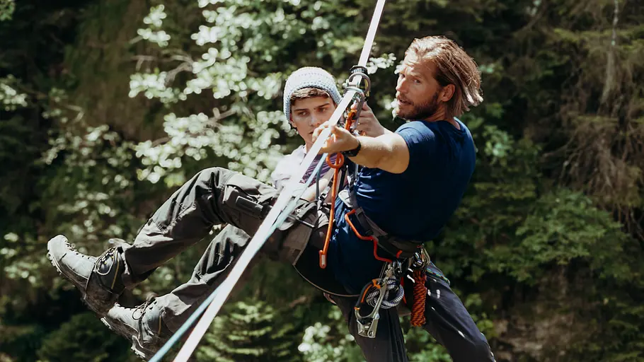 Jonas Meier (Juls Seger, hinten) und Markus Kofler (Sebastian Ströbel, r.) Die Bergretter: Ohne Ausweg: Im Wald: Über einem Abgrund ist eine Slackline gespannt. An dieser hängt Jonas Maier (Juls Serger) in seinem gesicherten Klettergurt und wirkt benommen. Vor ihm, ebenfalls gesichert, hängt Markus Kofler (Sebastian Ströbel) und zieht sich und Jonas an der Slackline entlang.