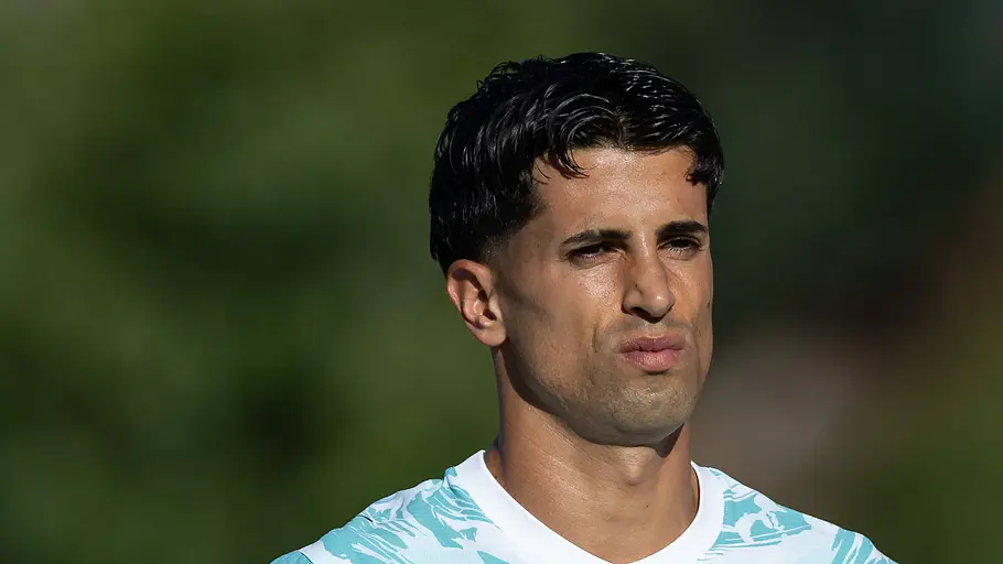 Fußballspieler im weiß-türkisen Trikot blickt mit konzentriertem Gesichtsausdruck in die Ferne. Im Hintergrund sind unscharfe grüne Bäume zu sehen.