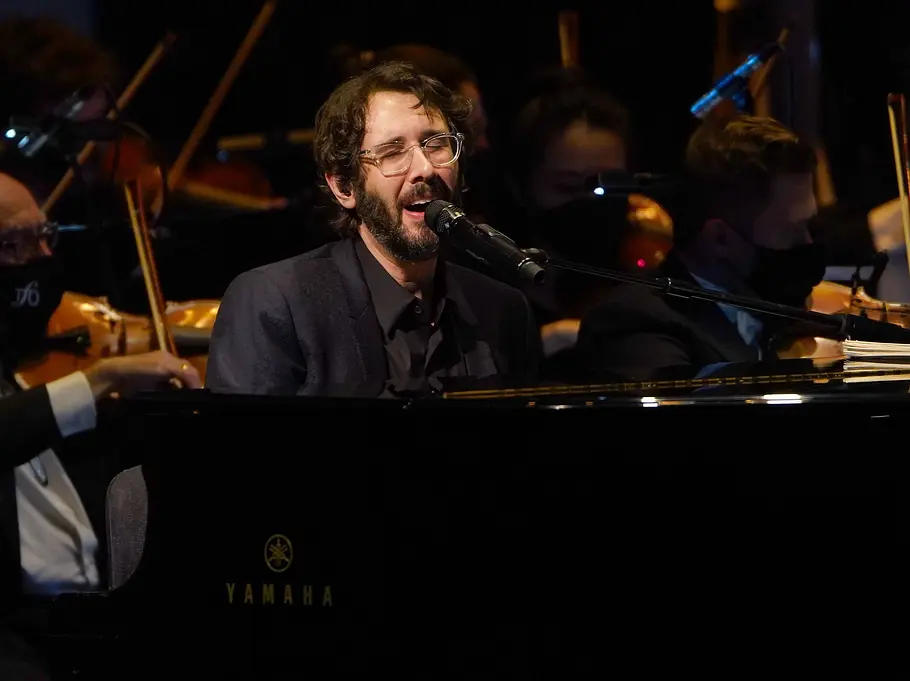 Josh Groban sitzt am schwarzen Flügel und singt ins Mikrofon. Hinter ihm spielen Orchestermusiker mit Gesichtsmasken.