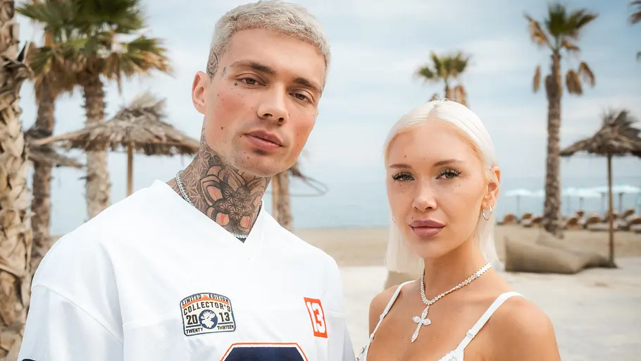 Josua Günther und Larissa sind seit 2025 ein Paar. Josua im weißen Trikot und Larissa im weißen Oberteil stehen vor Palmen und Sonnenschirmen am Strand.