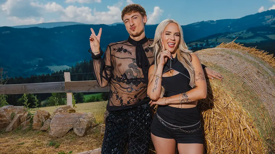 Gina Beckmann und Leon Content posieren nebeneinander vor einer Heuballenrolle in einer ländlichen, bergigen Landschaft. Leon trägt ein durchsichtiges, schwarzes, besticktes Oberteil und macht ein Peace-Zeichen, während Gina ein schwarzes Top und Shorts trägt und lächelt.