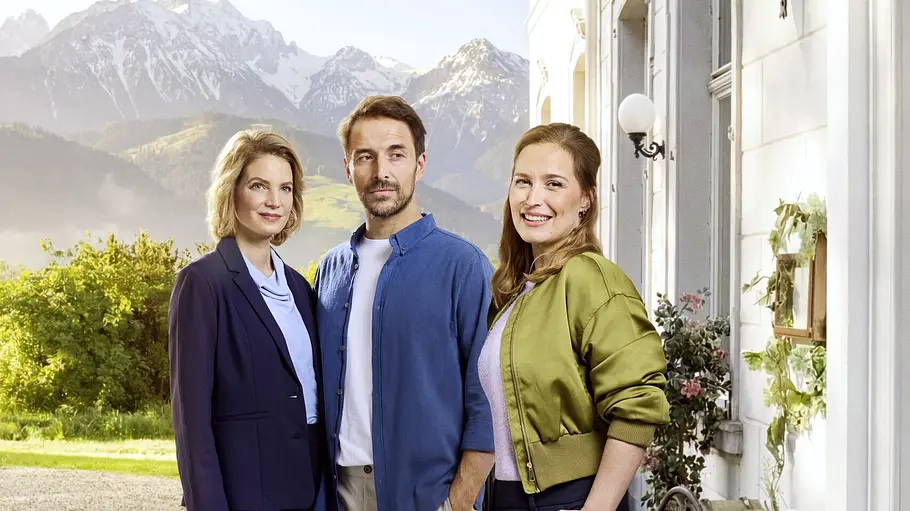 Stars von „Die Landarztpraxis“-team Sonnenhof Zwei Frauen und ein Mann stehen vor einem weißen Haus mit dem Schild „Sonnenhof“. Im Hintergrund sind Berge zu sehen.
