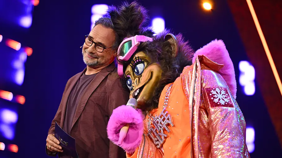 Matthias Opdenhövel Three Sixty Masked Singer Matthias Opdenhövel mit Brille steht auf einer Bühne neben Three Sixty im pelzigen Tierkostüm mit orange-pinker Glitzerjacke und Mikrofon.