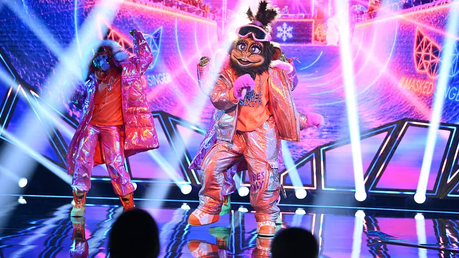 Figuren mit Affenmasken und glänzenden Outfits stehen auf einer bunt beleuchteten Bühne. Die vordere, Three Sixty, hält ein Mikrofon.