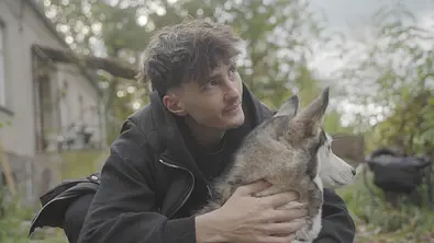 Ein junger Mann in dunkler Jacke kniet im Freien und hält einen Husky im Arm, während beide aufmerksam in die Ferne blicken. - Foto: Joyn/ProSieben