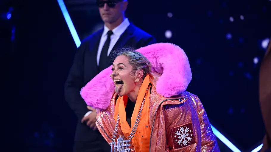 Ruth Moschner Masked Singer enthüllt Ruth Moschner in glänzender Jacke mit pinkem Fellkragen streckt die Zunge heraus. Dahinter steht ein Mann im Anzug mit Sonnenbrille.