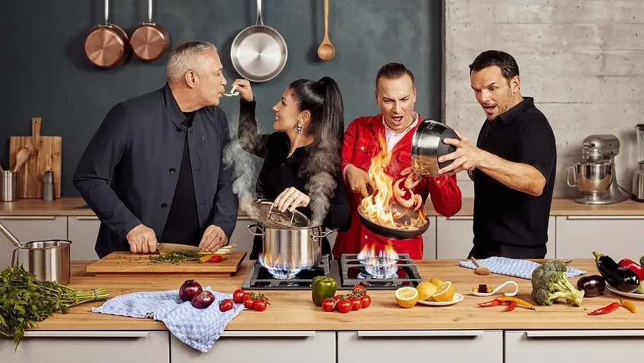 The Taste Staffel 15 Die The Taste-Jury steht in einer modernen Studio-Küche. Sie probieren ein Gericht, während in einer Pfanne auf dem Herd hohe Flammen schlagen.