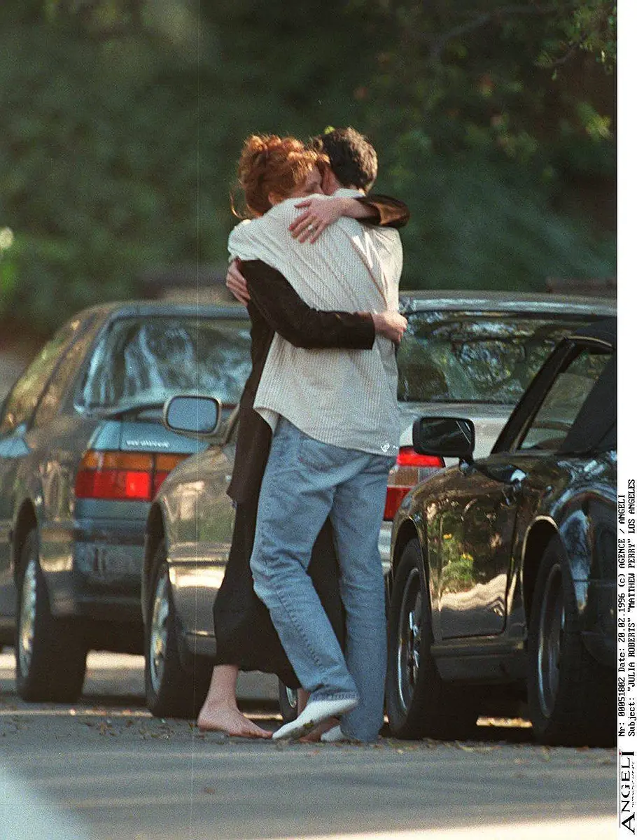 Julia Roberts und Matthew Perry umarmen sich zwischen geparkten Autos. Sie ist barfuß, trägt ein dunkles Kleid, er in Socken, hellem Hemd und Jeans.