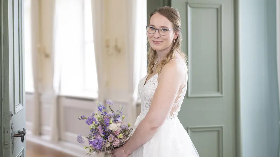 Julia Hochzeit auf den ersten Blick Julia trägt ein weißes Brautkleid. Sie hat einen Blumenstrauß mit lila, rosa, und weißen Blumen in den Händen.