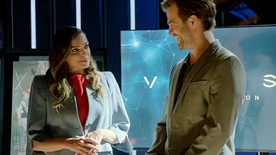 Emily und Julian stehen lächelnd voreinander. Sie trägt einen grauen Blazer über einem roten Oberteil, er ein graues Sakko. - Foto: RTL