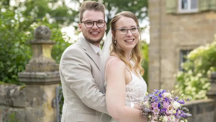 Ein frisch verheiratetes Paar steht lächelnd im Freien. Der Bräutigam in hellem Anzug umarmt die Braut, die ein weißes Spitzenkleid und einen bunten Blumenstrauß trägt. Im Hintergrund sind Grünpflanzen und ein historisches Gebäude zu sehen.