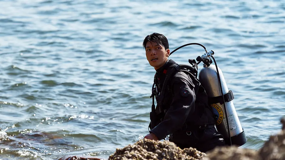 Jun-ho im Taucheranzug und mit Sauerstoffflasche auf dem Rücken. Er steht im Wasser, nah an der Küste.