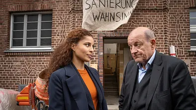 Wilsberg - Mogelpackung: Wilsberg (Leonard Lansink) und Tessa Tilker (Patricia Meeden) stehen vor einem Mehrfamilienhaus, das sich im Umbau befindet. Vor dem Gebäude stehen Baumaschinen, und aus einem Fenster hängt ein Banner mit der Aufschrift Sanierungswahnsinn. - Foto: ZDF/Thomas Kost Wilsberg - Mogelpackung: Wilsberg (Leonard Lansink) und Tessa Tilker (Patricia Meeden) stehen vor einem Mehrfamilienhaus, das sich im Umbau befindet. Vor dem Gebäude stehen Baumaschinen, und aus einem Fenster hängt ein Banner mit der Aufschrift Sanierungswahnsinn. - Foto: ZDF/Thomas Kost