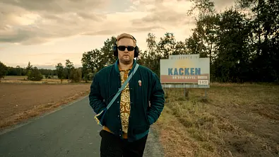 Anton Fatoni Schneider trägt eine Sonnenbrille, hat Kopfhörer auf und läuft gerade aus dem Ort Kacken an der Havel. Hinter ihm sieht man das Ortsschild. - Foto: Netflix