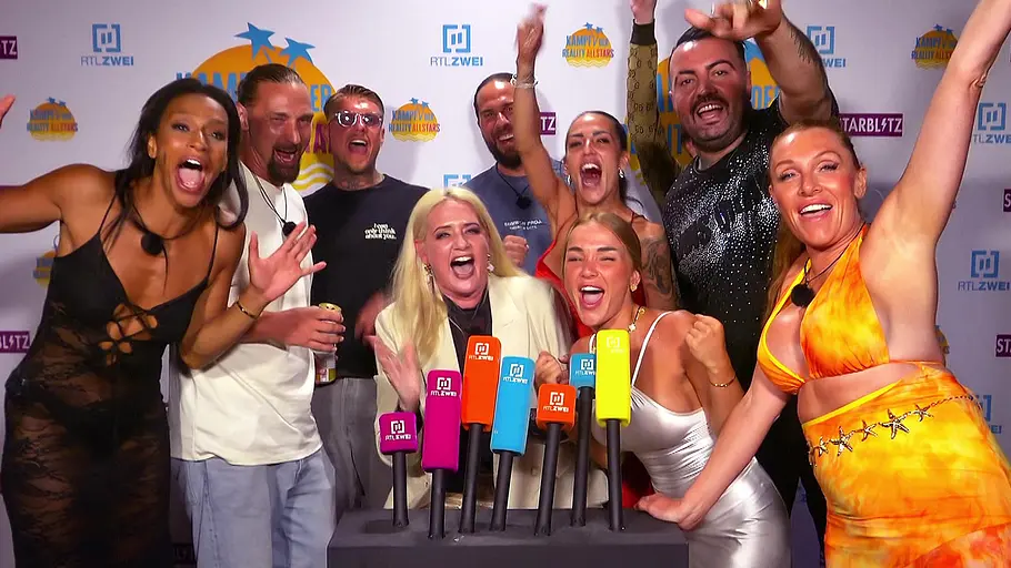 KDRS Eine Gruppe von Menschen steht dicht gedrängt vor mehreren bunten Mikrofonen, jubelt laut, reißt die Arme in die Höhe und wirkt ausgelassen wie bei einer TV-Show oder Pressewand mit Logos im Hintergrund.