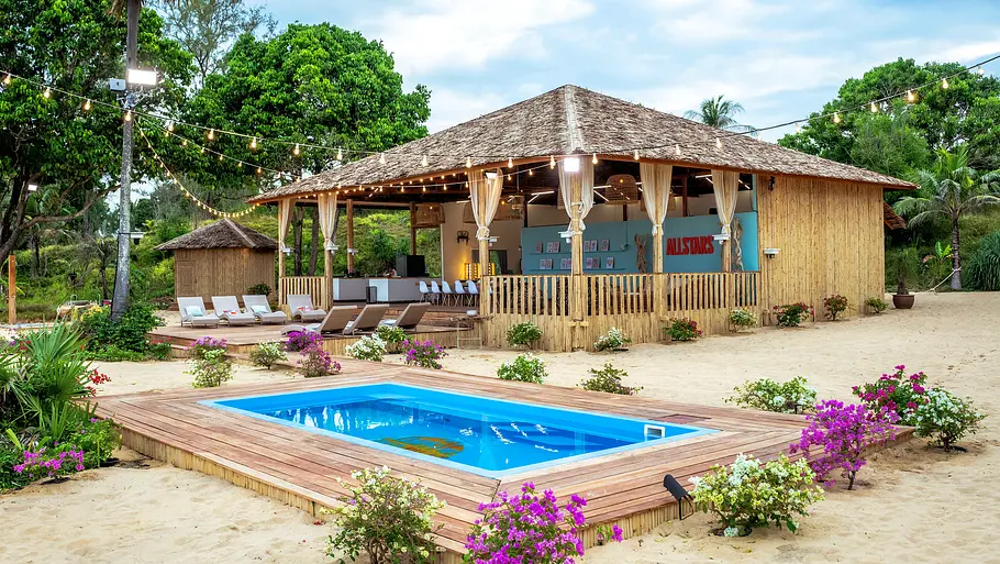KDRS Sala Tropische Strandkulisse mit einer offenen, strohgedeckten Holzhütte („Sala“) auf Sandboden. Vor der Hütte befindet sich ein rechteckiger Pool mit Holzterrasse, umgeben von pink blühenden Pflanzen und Palmen. Lichterketten spannen sich über das Gelände, neben der Terrasse stehen mehrere Liegen. An der Rückwand der Hütte ist ein „Allstars“-Schriftzug zu sehen.