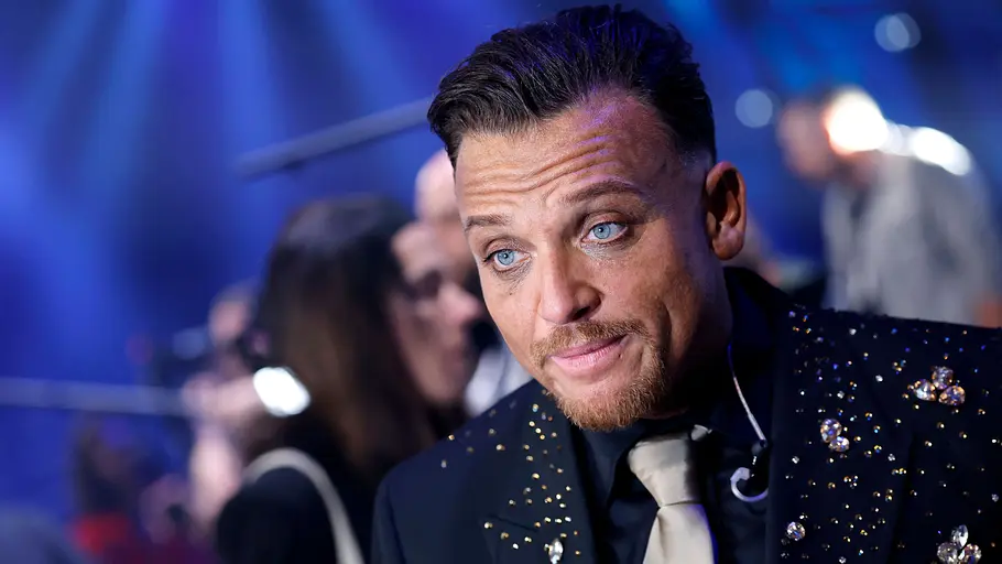 Menowin Fröhlich: Sidos Instagram-Post sorgt für Spekulationen Menowin Fröhlich in schwarzem, glitzerndem Sakko und heller Krawatte bei einem DSDS-Auftritt vor blau beleuchteter Bühne.