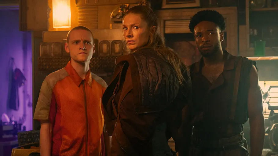 Lewis Gribben, Hunter Schafer und Dimitri Abold stehen in einem dämmrig beleuchteten Raum und blicken ernst nach vorn.