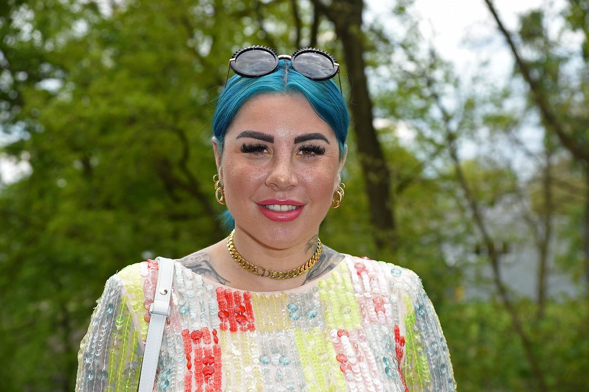 Kate Merlan Kater Merlan mit blauen Haaren und einer Sonnebrille auf dem Kopf trägt einen bunten Pullover und lächelt in die Kamera