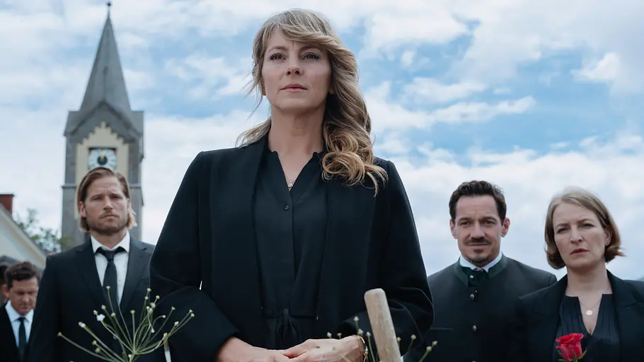 Katharina steht in schwarzer Kleidung vor einem Blumengesteck. Hinter ihr stehen Markus, Tobi und seine Frau, rechts mit Rosen, und ein Kirchturm.